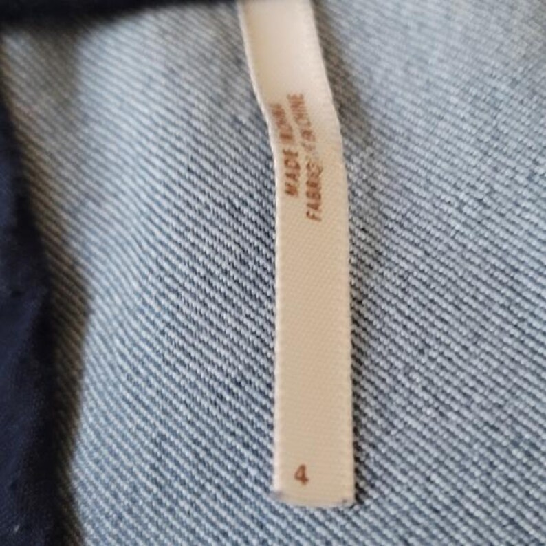 Puede incluir: Primer plano de una etiqueta de ropa color crema con el texto "MADE IN CHINA FABRIQU&Eacute; EN CHINE" y el n&uacute;mero "4". La etiqueta est&aacute; adherida a una tela vaquera azul.