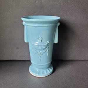 Może przedstawiać: Jasnoniebieski ceramiczny wazon z dekoracyjnym wzorem. Wazon posiada rozszerzoną krawędź, dwa boczne uchwyty oraz szczegółowy wytłoczony wzór misy i koralikowych girland. Wazon stoi na okrągłej podstawie.