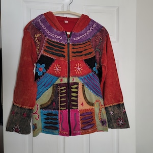 Peut inclure: Veste patchwork colorée avec capuche et fermeture éclair intégrale. La veste présente une variété de couleurs, dont le rouge, le violet, le bleu et le vert, avec des motifs floraux et étoilés brodés. Les manches ont des poignets évasés.