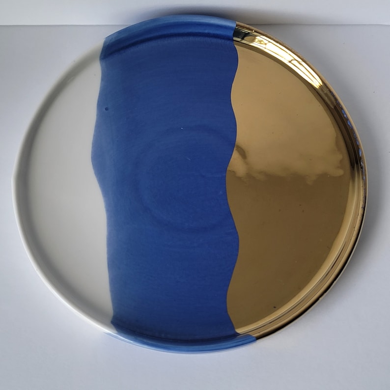 Puede incluir: Un plato redondo de cer&aacute;mica con fondo blanco y un dise&ntilde;o azul y dorado. El dise&ntilde;o azul es una l&iacute;nea ondulada que cubre la mitad del plato. El dise&ntilde;o dorado es un color s&oacute;lido que cubre la otra mitad del plato.