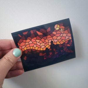 May include: A stained glass style sticker with a sunset scene and two figures silhouetted in the foreground. The sunset is a mix of orange, red, and yellow colors. The background is dark blue.