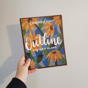 May include: A spiral bound book with a brown cover featuring a stained glass design of sunflowers and the text "Outline Stained Glass" and "CreationbyCarson".