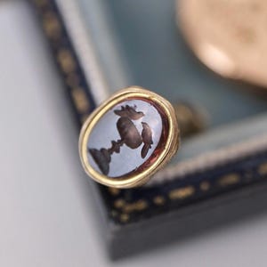 May include: A gold ring with an oval, pale blue stone featuring a dark brown, detailed design. The ring is set in a gold band and is displayed on a blue and gold box, with other gold items in the background.