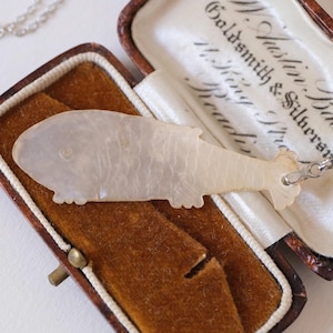 以下が含まれることがあります： 茶色のベルベットの裏地が付いた箱に入った、ヴィンテージの魚のペンダントネックレス。ペンダントは真珠光沢があり、シルバーチェーンが付いています。箱は開いており、ペンダントとチェーンが見えます。箱には金の留め金が付いています。