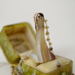May include: An open, antique-style green jewelry box with a cream-colored interior holds a decorative, brown and white stone pendant. A delicate necklace with small, green beads drapes from the pendant. The box is closed with a small metal clasp.