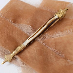 May include: An antique letter opener with a gold-coloured handle and a bone-coloured shaft with brown marbling. The handle is shaped like a pointing hand. The letter opener rests on a peach-coloured ribbon.