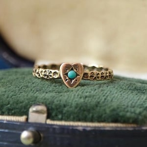 Puede incluir: Un anillo de oro con una pieza central en forma de corazón que tiene una pequeña piedra turquesa redonda de color azul brillante engastada en el centro. La banda del anillo presenta un patrón geométrico texturizado que rodea toda la circunferencia.