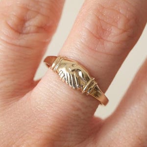 May include: A gold-colored ring featuring a detailed design of two clasped hands. The ring is worn on a finger, showcasing its intricate craftsmanship and warm metallic hue. The ring's design symbolizes unity and agreement.