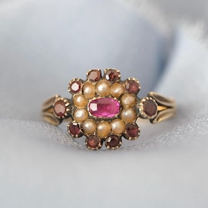 Puede incluir: Un anillo de oro con una gema rosa ovalada central, rodeada de pequeñas perlas y gemas rojas. El diseño del anillo incluye gemas rojas adicionales en la banda, creando un patrón floral. El anillo se encuentra sobre una tela azul claro.