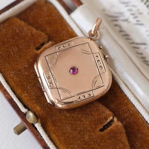 May include: A gold-colored, square locket with a small, round, red gemstone in the center. The locket has etched geometric designs and is displayed open inside a brown velvet-lined case with a leather exterior. The case is open, revealing the locket.
