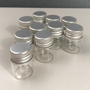 May include: A group of 12 small, clear glass bottles with silver lids. The bottles are arranged in a semi-circle.