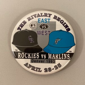 May include: A white button with a black and blue baseball cap design. The text on the button reads "The Rivalry Begins", "East vs West", "Rockies vs Marlins", "Denver 1993", and "April 23-25".