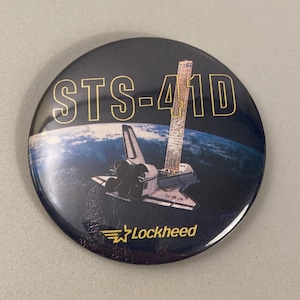 May include: A round button with a black background and a gold inscription that reads "STS-41D". The button features a space shuttle in flight over a blue and white Earth. The Lockheed logo is visible in the lower right corner.
