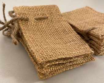 May include: Several small, rectangular burlap bags are stacked and arranged on a light gray surface. Each bag has a drawstring closure made of twine. The bags are a natural tan color and have a coarse, woven texture.