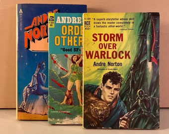 May include: Three vintage science fiction paperback books with colorful covers. The book on the left has a blue cover with a white and blue illustration of a creature. The middle book has a yellow cover with a woman in a white and blue outfit. The book on the right has a blue cover with a man in a blue jacket and a creature with brown fur. The text on the covers reads "Andre Norton", "Storm Over Warlock", "Good SF", "Order Other", and "Andre Norton".