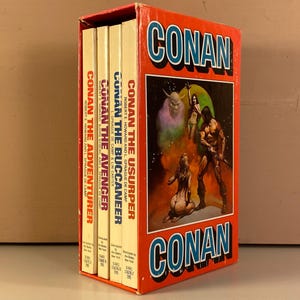 May include: A boxed set of four vintage Conan the Barbarian paperback books. The box and book spines are red with white text. The box features a fantasy illustration of Conan and other figures. Titles include "The Adventurer", "The Avenger", "The Buccaneer", and "The Usurper."