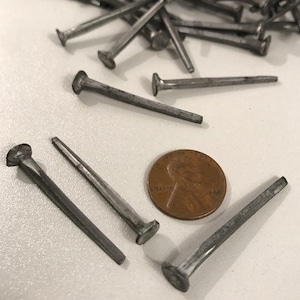 May include: A close-up of a pile of silver-colored tacks or nails with a penny for scale. The tacks are about 1 inch long and have a flat head.