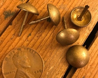 Può includere: Cinque chiodini antichi color oro con teste rotonde e un gambo a forma di chiodo. I chiodini sono disposti su una superficie di legno.
