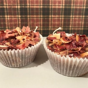 May include: Two small, cupcake-shaped candles filled with reddish-brown wood shavings. The candles are in white paper liners and have white wicks. The background is a plaid pattern in shades of brown and red.