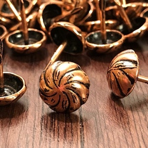 May include: A close-up of a pile of copper-colored tacks with black centers and a swirl design. The tacks have a nail-like shaft and are scattered on a wooden surface.