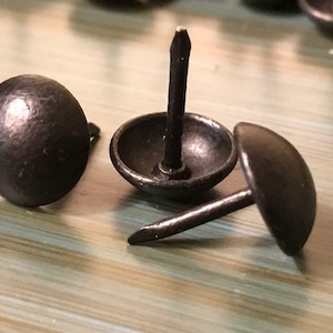 May include: Three black metal tacks with round heads and sharp points. The tacks are on a wooden surface.