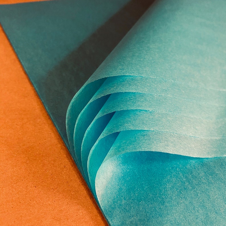 May include: Close-up of several sheets of teal tissue paper, fanned out to reveal the layers. The paper has a soft, translucent quality, with subtle variations in color. The background is a warm, earthy orange.