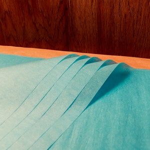 May include: A stack of folded, light blue tissue paper sheets. The paper is arranged in a wave-like pattern, with each sheet slightly overlapping the next. The background features a wooden surface.
