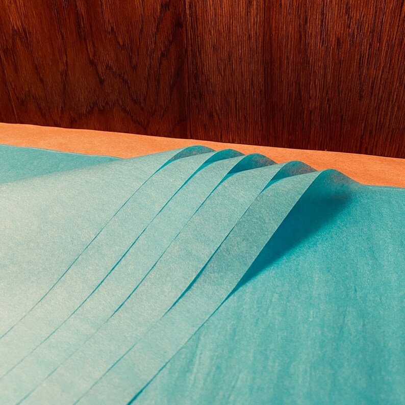 May include: A stack of folded, light blue tissue paper sheets. The paper is arranged in a wave-like pattern, with each sheet slightly overlapping the next. The background features a wooden surface.