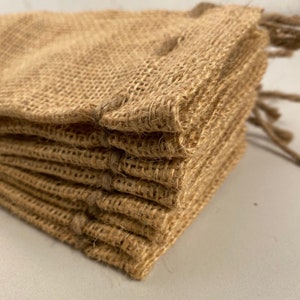 May include: Close-up of a stack of small, beige burlap bags. The bags are made of a coarse, woven fabric and have a natural, rustic appearance. Each bag is folded over at the top, and a twisted jute string is visible.