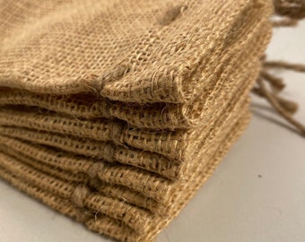 May include: Close-up of a stack of small, beige burlap bags. The bags are made of a coarse, woven fabric and have a natural, rustic appearance. Each bag is folded over at the top, and a twisted jute string is visible.