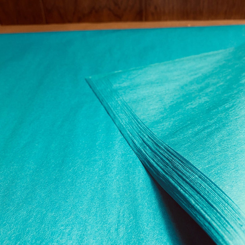 May include: A stack of teal-colored tissue paper, with a slightly textured surface. The paper is neatly stacked, with the edges forming a sharp corner. The background is a wooden surface.