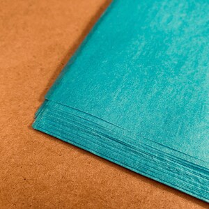 May include: A stack of turquoise tissue paper sheets. The paper has a textured surface and is arranged on a brown background. The corner of the stack is visible, showing the layered edges of the paper.