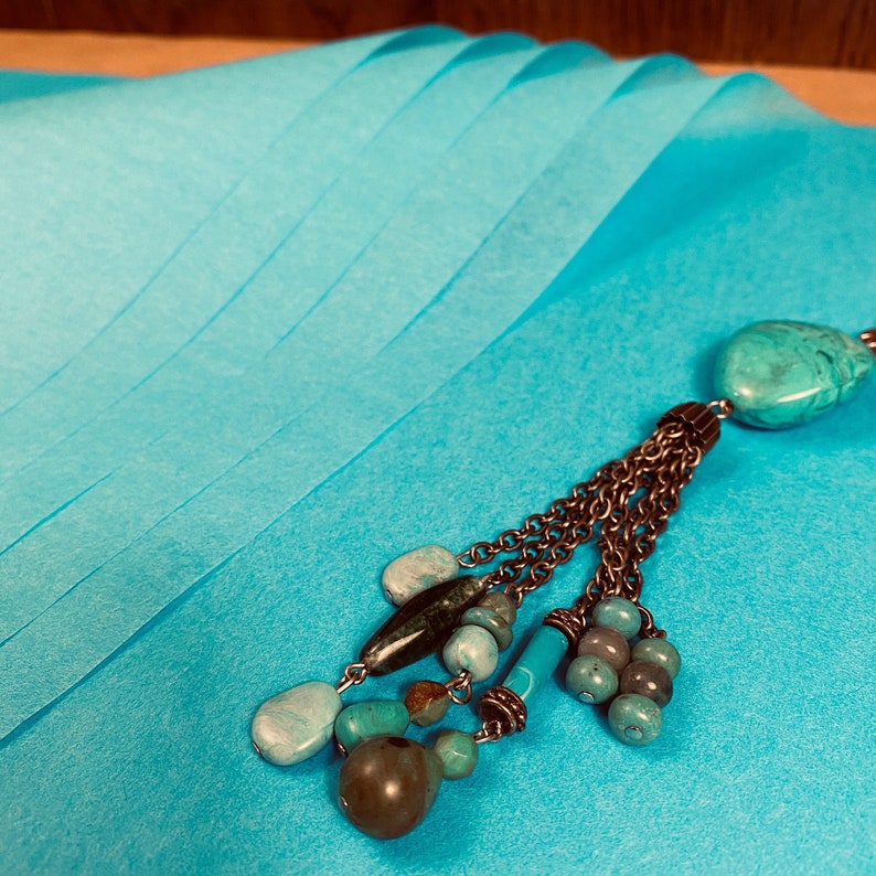 May include: A turquoise pendant necklace with a teardrop-shaped stone and a chain tassel design. The necklace features various turquoise and brown beads. The background is a folded, light blue paper.