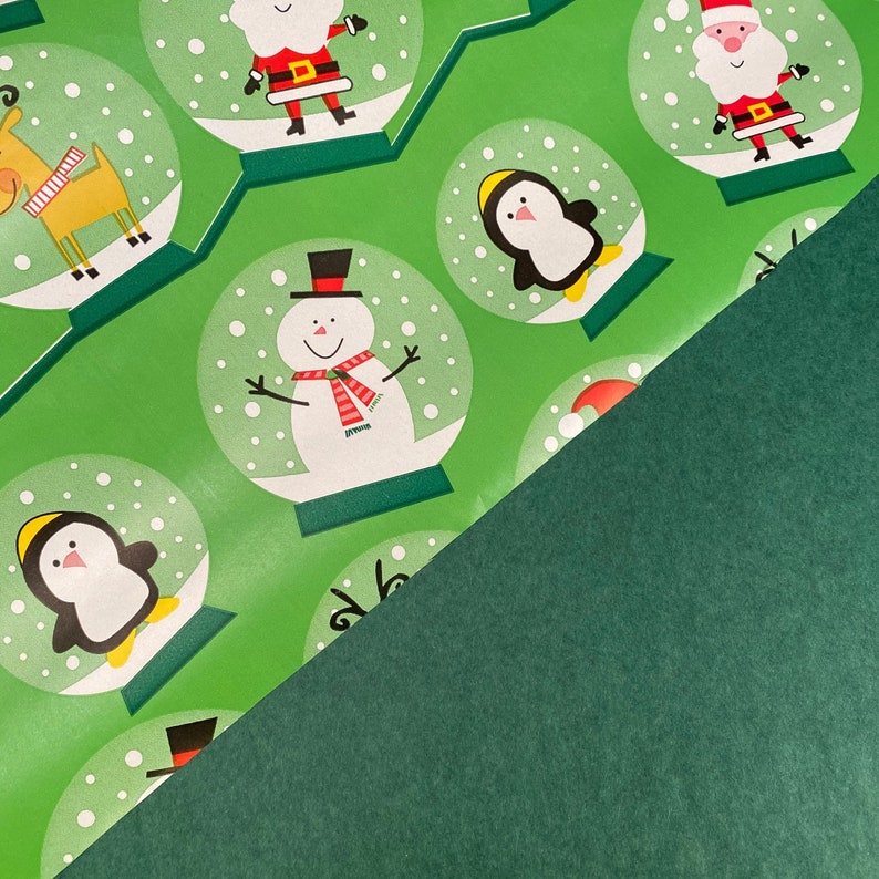 Puede incluir: Papel de regalo verde con un patr&oacute;n repetido de bolas de nieve. Cada bola presenta un personaje navide&ntilde;o diferente, incluyendo un mu&ntilde;eco de nieve, un ping&uuml;ino, un reno y Santa Claus. El papel est&aacute; parcialmente cubierto por una hoja verde s&oacute;lida.
