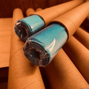 May include: Several rolls of turquoise paper are visible, each encased in a brown cardboard tube and wrapped in clear plastic. The tubes are arranged at various angles, creating a layered effect. The paper appears to be a solid color.