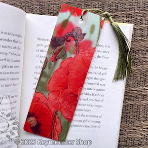 May include: A rectangular bookmark featuring a vibrant design of red poppies and a small gray cat. The bookmark has a green tassel and is placed in an open book. The text "© 2025 Keymonster Shop" is visible.