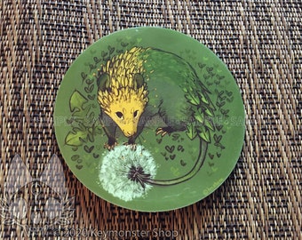 May include: A round sticker featuring a green and yellow possum with a dandelion in front of it. The possum has a spiky back and a long tail. The background is green with small white hearts and green leaves.