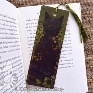 May include: A bookmark featuring a black cat illustration surrounded by green leaves. The bookmark has a green tassel and is placed in an open book. The words "SAMPLE" are printed on the bookmark.