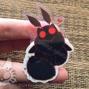 May include: A clear sticker with a black and red moth design. The moth has large, red eyes and a heart-shaped design above it. The sticker is on a brown, woven surface.