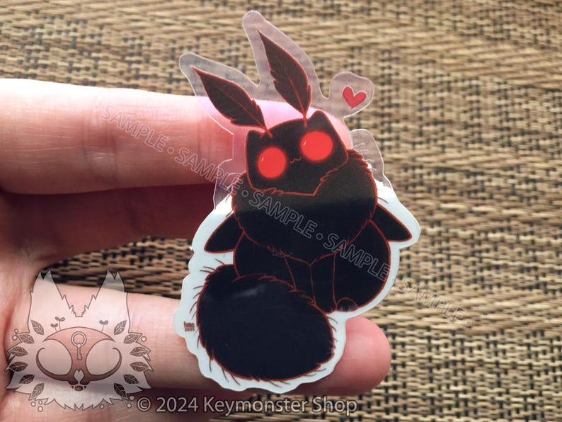 May include: A clear sticker with a black and red moth design. The moth has large, red eyes and a heart-shaped design above it. The sticker is on a brown, woven surface.