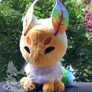 May include: A plush toy creature with a white body, yellow face, and rainbow-coloured leaf-shaped ears. The toy has a fluffy white collar and a rainbow tail. The text "© 2025 Keymonster Shop" is visible.