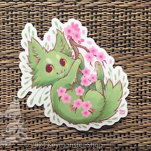 May include: A green cat-like creature with pink flowers and leaves. The creature is holding a branch with pink flowers. The background is a woven brown mat.