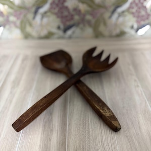 May include: A pair of crossed wooden salad servers. One server is a spoon, the other a fork with three tines. The servers are a dark brown color and appear to be made of a solid wood. The background is a floral pattern.