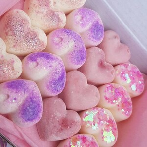 May include: A box filled with heart-shaped wax melts in various shades of pink, purple, and white. Some melts have glitter embedded in them. The box is labeled "Kimmy's Scents".