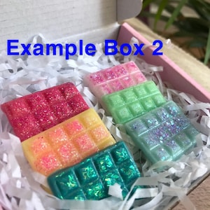 May include: A box filled with white shredded paper containing six wax melts. The wax melts are in a variety of colors, including pink, green, yellow, and blue. The wax melts are all square shaped and have a glittery finish. The box has the text "Example Box 2" printed on the top.