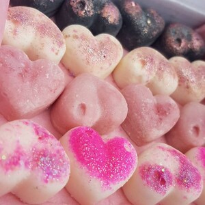 May include: A box of heart-shaped wax melts in various shades of pink, white, and black. Some hearts are dusted with glitter. The box has a pink interior and a label with the text "Kimmy's Scents".