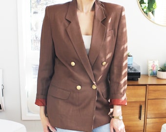 May include: A brown double-breasted blazer with gold buttons. The blazer has a slightly oversized fit and is made of a textured fabric. The blazer is worn over a white tank top and blue jeans.