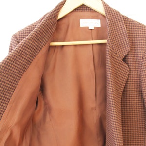 Può includere: Un blazer marrone e nero a pied-de-poule con fodera marrone. Il blazer è sbottonato e appeso a un appendiabiti di legno.