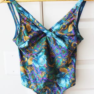 May include: A vintage one-piece swimsuit with a vibrant floral and paisley pattern in shades of blue, purple, and gold. The swimsuit features a V-neckline and thin straps, hanging on a wooden hanger.