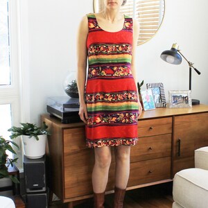 May include: A woman wearing a sleeveless red, green, yellow, and orange patterned dress with a scoop neck. She is standing in front of a wooden dresser with two drawers.